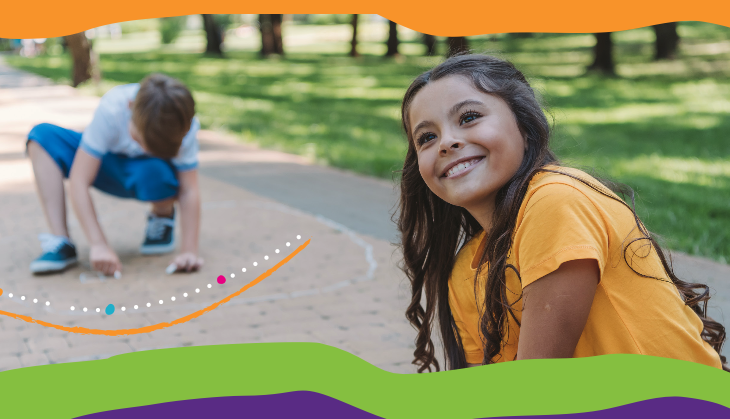 Smiling girl in yellow short in foreground and boy drawing on concrete with chalk in background