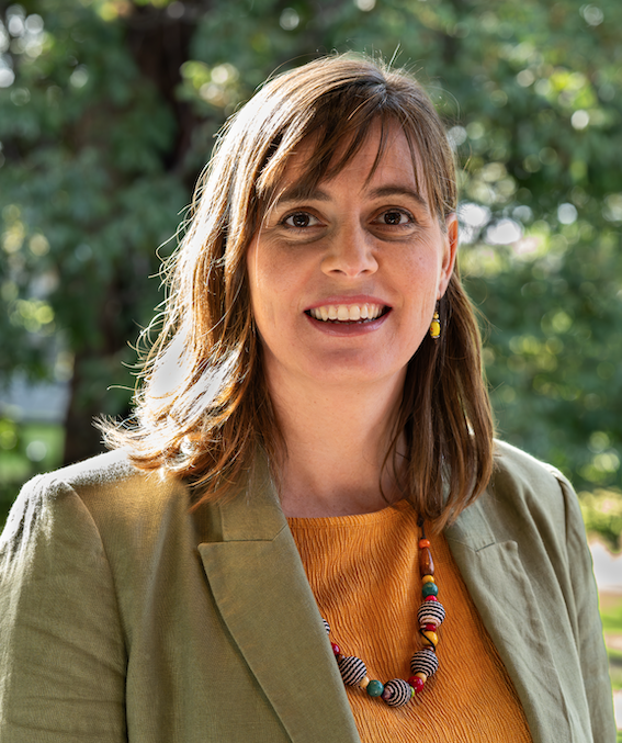 Woman with long brown hair, orange top, jacket and necklace, with treed background